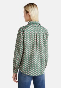 Camicia verde con un motivo floreale rosa, maniche lunghe, polsini con bottoni e una piega sul retro. Il tessuto sembra liscio con una vestibilità rilassata.