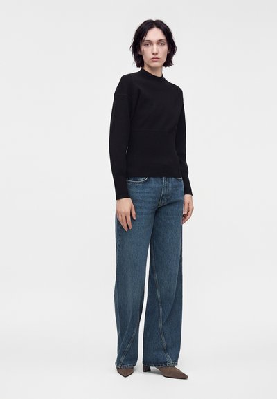 Γυναίκα με κοντά σκούρα μαλλιά, ντυμένη με στενό μαύρο μακρυμάνικο τοπ, φαρδιά μπλε τζιν και καφέ μυτερές παπούτσια, στέκεται μπροστά σε απλό φόντο.