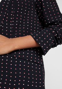 Black fabric top featuring a dotted pattern in red and white. The design includes long sleeves with flared cuffs and a relaxed fit.