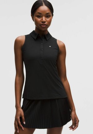 Žena v čiernom bezrukávovom topu s golierom a čiernej plisovanej sukni stojaca pred svetlým jednoliatym pozadím.