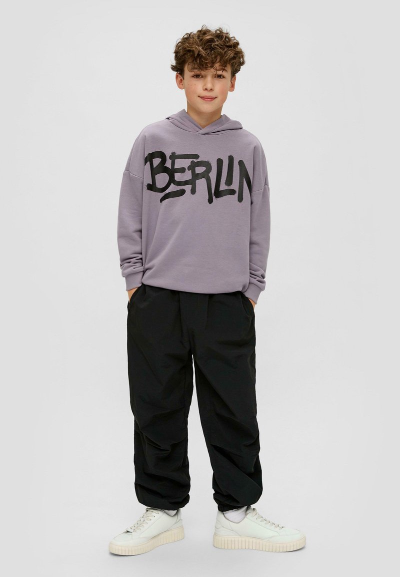 Paarse hoodie met "BERLIJN" in zwarte letters, gecombineerd met losse zwarte broek en witte high-top sneakers. Glad stof met een relaxte pasvorm.