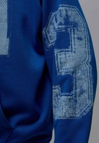 Niebieska bluza z dużą, fakturowaną cyfrą „3” w jasnoniebieskim kolorze na lewym rękawie, wykonana z gładkiej i miękkiej bawełny.