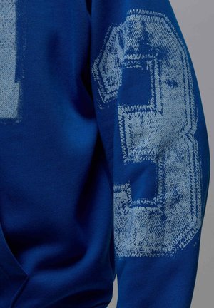 Niebieska bluza z dużą, fakturowaną cyfrą „3” w jasnoniebieskim kolorze na lewym rękawie, wykonana z gładkiej i miękkiej bawełny.