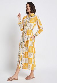Long-sleeve, button-up dress with a yellow and cream geometric pattern, featuring a belted waist and side pockets. Accompanied by pink sandals.