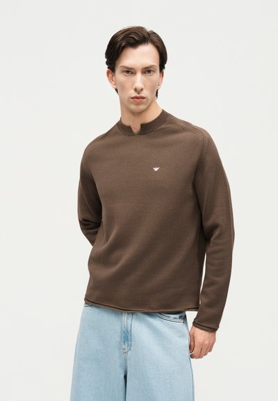 Άνδρας που φοράει καφέ πλεκτό πουλόβερ με μικρό λογότυπο στο στήθος και χαλαρά ανοιχτό μπλε τζιν, στέκεται μπροστά σε απλό φωτεινό φόντο.