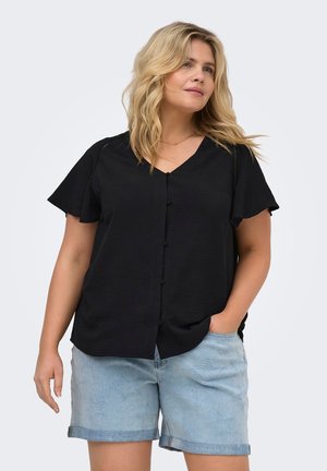 Femme aux cheveux blonds portant un chemisier noir à manches courtes boutonné et un short en jean bleu clair, une main dans la poche.