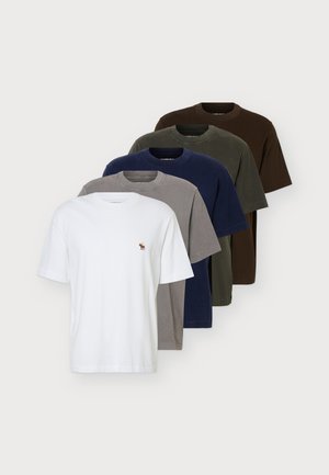 Un homme porte un tee-shirt bleu marine avec un petit logo sur la poitrine, un jean bleu clair et des chaussures marron sur un fond clair et uni.