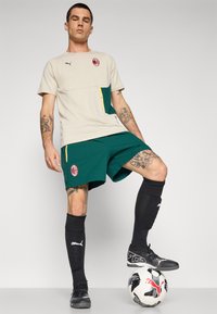Jasny beżowy sportowy podkoszulek z zielonymi akcentami, logo Puma i czerwonym herbem. Zielone szorty z bocznymi kieszeniami, czarne skarpetki sięgające do kolan i czarne korki na piłce nożnej.