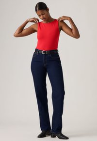 Γυναίκα που προσαρμόζει το κόκκινο αμάνικο τοπ, φοράει σκούρο μπλε τζιν με μαύρη ζώνη και μαύρα μπότες με τακούνι, στέκεται μπροστά σε απλό φωτεινό φόντο.