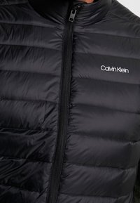 Czarna pikowana kamizelka puchowa z wysokim kołnierzem i zamkiem błyskawicznym na całej długości. Na lewej piersi znajduje się dyskretne logo Calvin Klein.