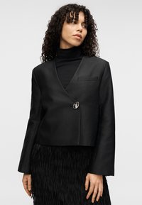 Μαύρο κοντό σακάκι με διαγώνια μπροστινή όψη, μονή ασημένια κλειστή λάμα, και μινιμαλιστική τσέπη. Φορεμένο πάνω από μαύρο ζιβάγκο και φούστα με κρόσσια.