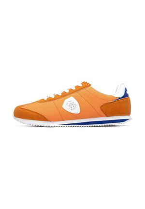 Zapatilla de tela y ante naranja con cordones blancos, un parche con el logo blanco en el costado y un detalle azul en el talón. Suela de goma con tracción.