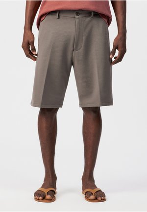 Mann trägt strukturierte graue knielange Shorts und beige Sandalen, steht vor einem schlichten weißen Hintergrund.