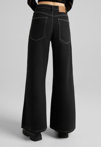 Breda svarta jeans med kontrasterande vit sömnad, bakfickor och en synlig läderetikett på midjan. Slen tygkänsla.