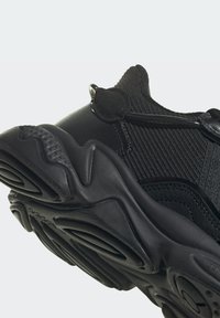 Černá sportovní bota s texturovaným síťovaným svrškem, zaoblenou gumovou podešví a elastickými tkaničkami, která zdůrazňuje aerodynamický design.