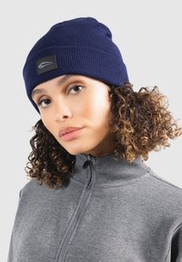 Beanie a maglia blu navy con una texture a coste e una toppa nera, abbinato a una giacca grigia con zip e colletto alto.