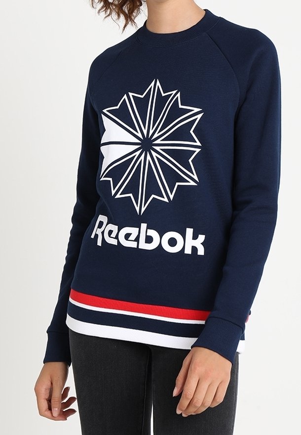 Námořnická modrá mikina s velkým bílým grafickým designem hvězdy a textem "Reebok". Obsahuje ribbovaný límec a pruhovaný lem v červené a bílé barvě.