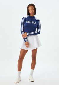 Chaqueta navy con cremallera y el texto "FRANCIA", con franjas blancas en las mangas. Combinada con una falda acampanada blanca y zapatillas blancas.