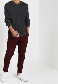 Mann in einem dunkelgrauen Strickpullover, burgunderfarbenen Hosen und weißen Sneakers, der vor einem schlichten weißen Hintergrund steht.