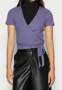 Lila rynkad wrap-top med korta ärmar, med en sidoknut. Bärs över en svart polotröja och matchas med svarta läderbyxor.