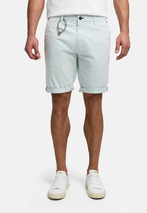 Męski model noszący jasnoniebieskie szorty z podwiniętym brzegiem oraz białe trampki VEJA, stojący na jednolitym białym tle.