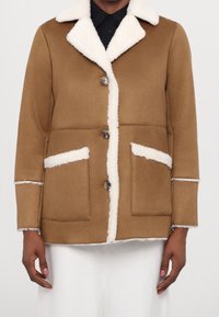 Hnědá bunda s ovčím límcem, bílými akcenty a dvěma předními kapsami. Obsahuje knoflíky na přední straně a texturovaný povrch.