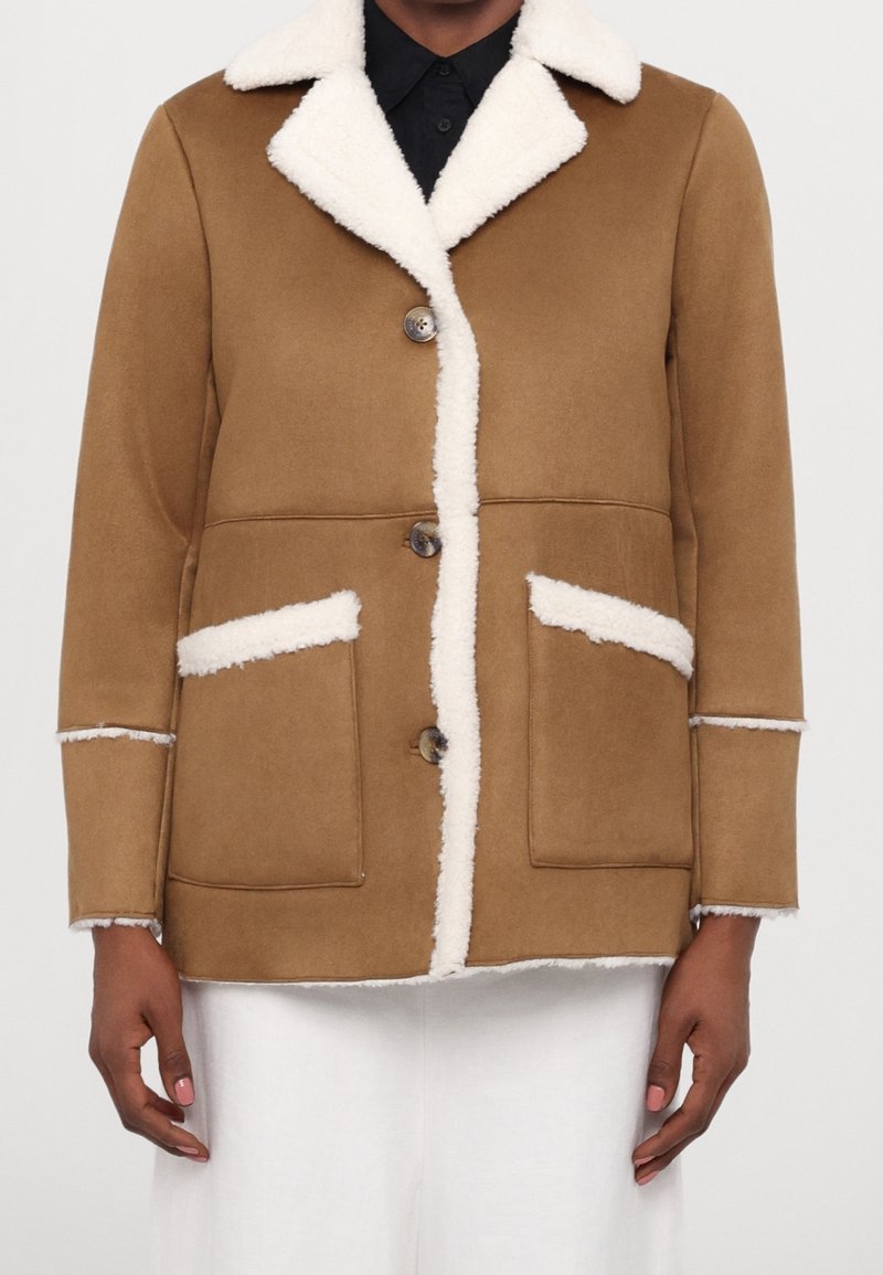 Hnědá bunda s ovčím límcem, bílými akcenty a dvěma předními kapsami. Obsahuje knoflíky na přední straně a texturovaný povrch.