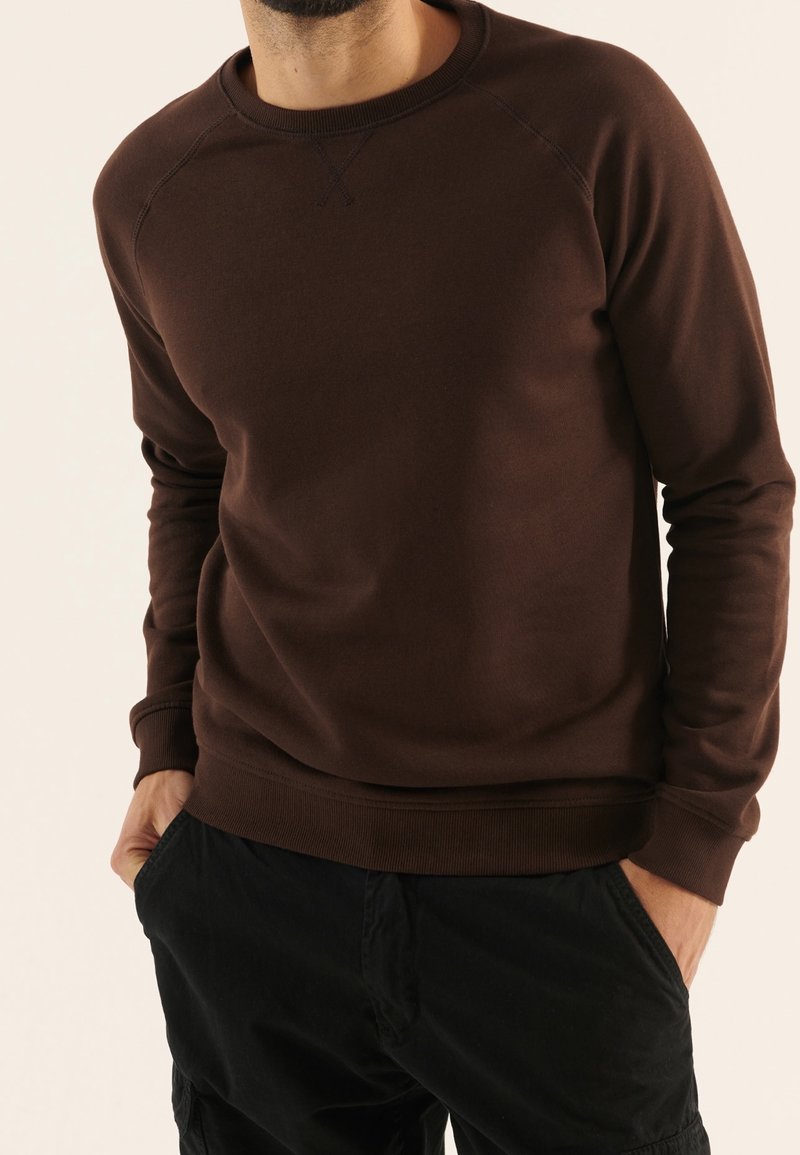Homme portant un sweat-shirt marron foncé à col rond avec manches longues et un pantalon noir, mains dans les poches.