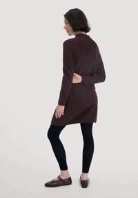 Langes, kastanienbraunes, geripptes Kleid, knielang, mit hohem Ausschnitt, langen Ärmeln, seitlichen Schlitzen, kombiniert mit schwarzen Leggings und braunen Schuhen.