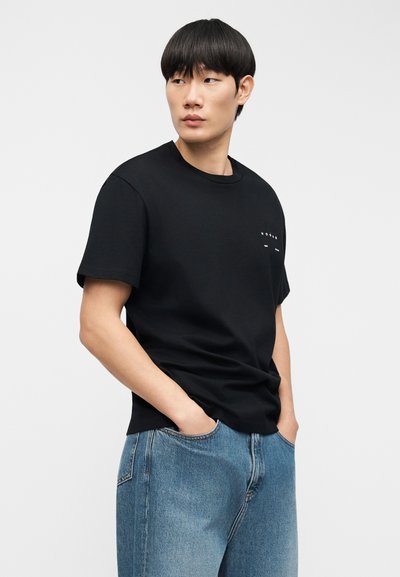 Μαύρο βαμβακερό μπλουζάκι με κοντά μανίκια, στρογγυλή λαιμόκοψη και μικρό λευκό λογότυπο στο στήθος, συνδυασμένο με ανοιχτόχρωμο μπλε τζιν παντελόνι.