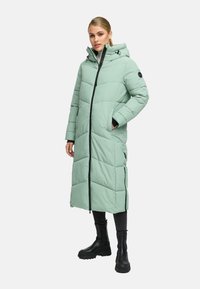 Woman wearing a long, quilted light green puffer coat with a hood, black zipper, side zippers, black pants, and chunky black boots.