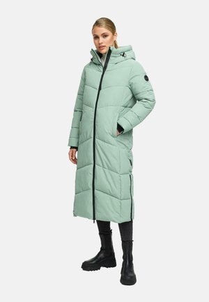 Kvinde iført en lang, quilted lys grøn pufferjakke med hætte, sort lynlås, sidelommer, sorte bukser og kraftige sorte støvler.