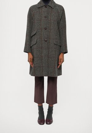 Manteau à carreaux marron mi-long avec de grands boutons et des poches à rabat, associé à un pantalon marron court, des chaussettes grises et des chaussures bordeaux.