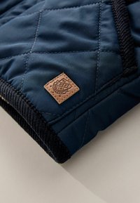 Quilted navy fabric with black ribbed hem, featuring a rectangular tan leather patch with embossed logo detail.