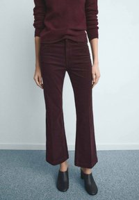 Persoană purtând pantaloni evazați de culoare burgundia închisă, pulover asortat și cizme negre cu toc până la gleznă, stând pe un covor gri.