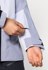 Svetlo siva jakna z dolgimi rokavi z črnimi detajli in zadrgo na rokavu. Gladek material z zasnovo prilegajočega se manšeta.