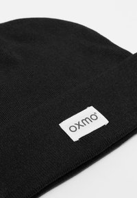Crna pletena kapa s preklopom, koja ima pravokutnu bijelu oznaku sa simbolom "oxmo" ušivenim u crnoj boji. Glatka tekstura i fleksibilno pristajanje.
