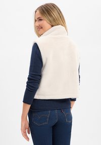 Cream faux fur vest with a high collar, paired with a navy long-sleeve shirt. Features a casual fit and soft texture.
