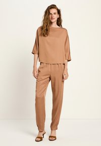 Frau mit langen, welligen Haaren, die ein lockeres braunes Seidentop, passende schmal geschnittene Hose und braune offene Heels trägt, steht vor einem neutralen Hintergrund.