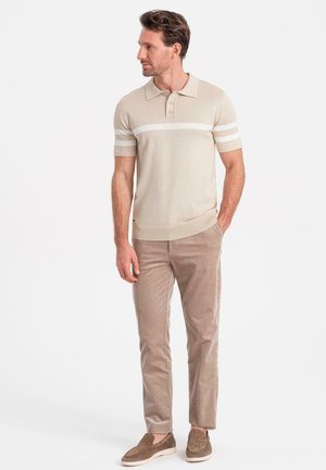 Beige Polo-Shirt mit weißen Streifen, kurzen Ärmeln und einem gerippten Kragen, kombiniert mit hellbraunen Cordhosen und braunen Slippern.