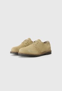 Scarpe in suede beige con lacci, con punta arrotondata, superficie texturizzata e suole in gomma scura a contrasto. Presentano cuciture sottili e un design semplice.
