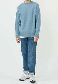 Maglione azzurro chiaro a maglia con bordi a coste, indossato sopra una camicia bianca, abbinato a jeans blu sbiaditi e sneaker bianche.
