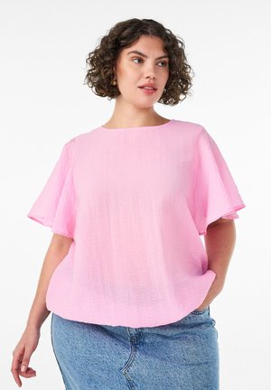 Mujer con cabello corto y rizado que lleva una camiseta suelta de manga corta rosa claro y jeans azules, de pie con una mano en el bolsillo sobre fondo blanco.