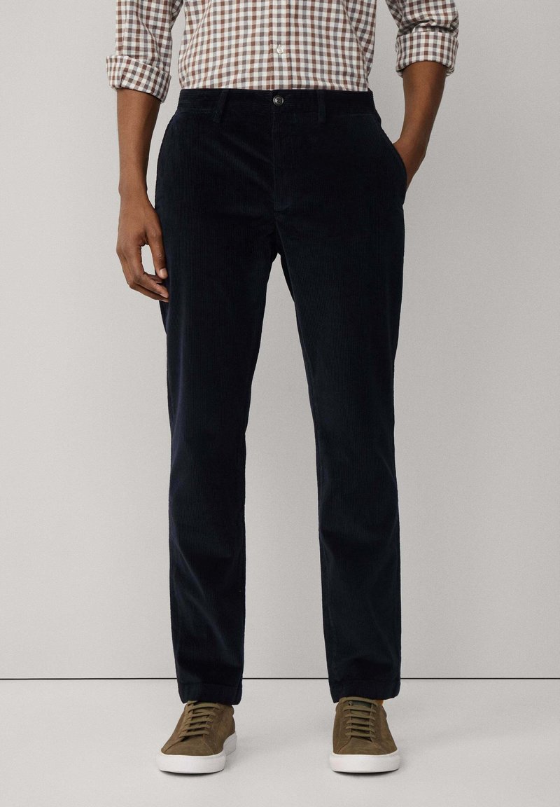Homme portant un pantalon en velours côtelé bleu marine foncé, une chemise à carreaux marron avec les manches retroussées, et des baskets marron avec des semelles blanches.