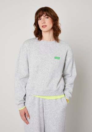 Õlgadeni pruunide juustega naine, kes kannab helehallist dressipluusi ja sobivaid pükse, seisab tavalise tausta ees.