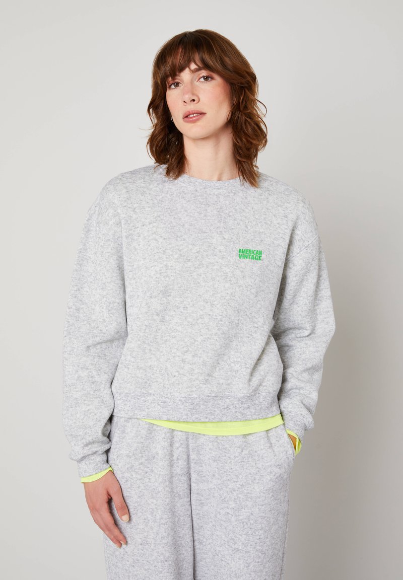 Kobieta z brązowymi włosami do ramion, ubrana w jasnoszarą bluzę i pasujące spodnie, stojąca na jednolitym tle.