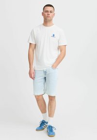 Vit bomull t-shirt med logotyp, ljusblå denimshorts och blå sneakers med snören. Modell står med händerna i fickorna.