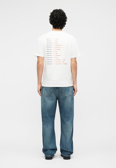 Άτομο με σγουρά μαλλιά που φοράει λευκό μπλουζάκι με πολύχρωμο κείμενο στο πίσω μέρος και φαρδιά μπλε τζιν, στέκεται μπροστά σε λευκό φόντο.