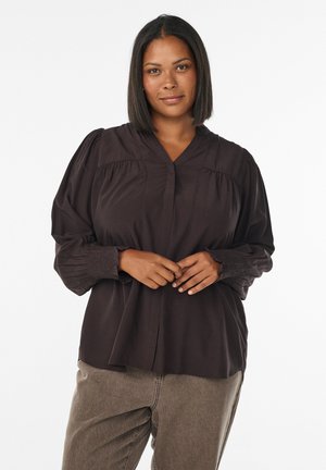 Vrouw met steil donker haar, gekleed in een zwarte blouse met lange mouwen en een bruine broek, staand met gekruiste handen tegen een witte achtergrond.