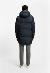 Cappotto puffer blu navy con cappuccio, caratterizzato da un design trapuntato e di lunghezza lunga. Abbinato a pantaloni grigi a gamba affusolata e sneakers bianche.